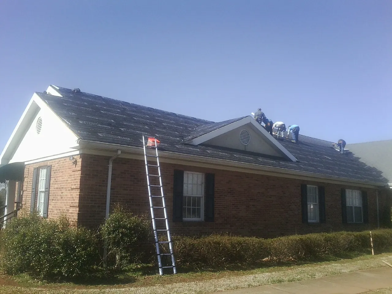 Crew tearing shingles off a brick ranch roof with a ladder set — Metal Roof Repair work by our crew in Rogers